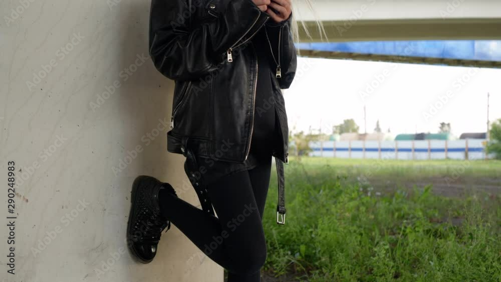 Rock girl in black leather jacket using mobile phone outdoor. Punk girl looking smartphone under car bridge in city. Urban life modern youth culture