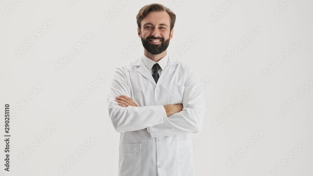Smiling young bearded man doctor in white professional coat winking and pointing with finger to the camera over gray background isolated