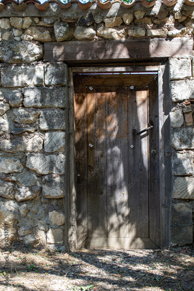 An old door with a rusty handle in a natural stone fence and a tiled roof. Treat sun falling through a big tree. An old door with a rusty handle in a natural stone fence and a tiled roof. Treat sun falling through a big tree.
