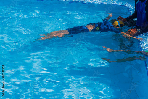 Asian little boy age 5 years old learning basic to swim in the lesson of lying and floating technique on surface water with adult male swimmer and trainer in blue swimming pool background.