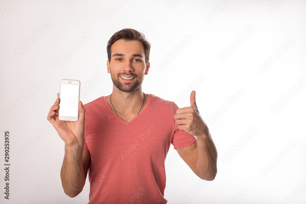 Smiling cheerful caucasian man in pink t-shirt showing hand sign well and holding mobil phone with empty mock up copy space screen background for promotional content, standing isolated in studio