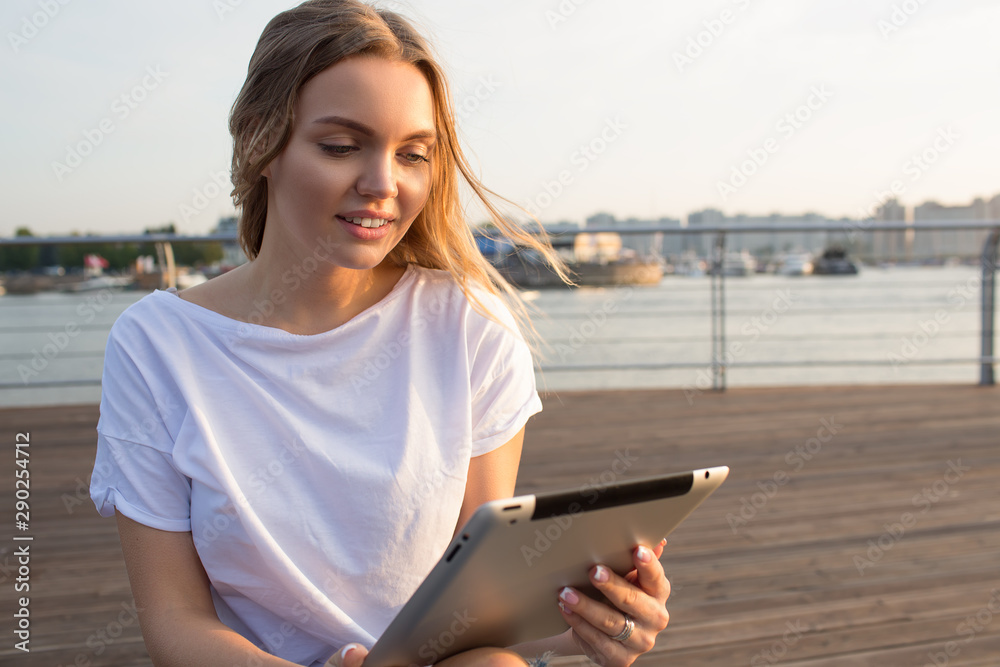 Gorgeous smiling woman content writer reading article on website via portable touch pad while relaxing on embankment during recreation time in summer weekend. Female online ordering via digital tablet