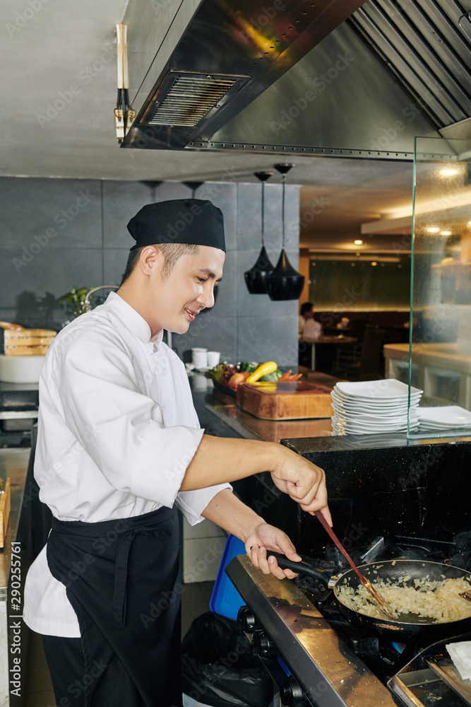 Asian young chef in uniform standing near the stove and frying rice on the pan in the kitchen Asian young chef in uniform standing near the stove and frying rice on the pan in the kitchen