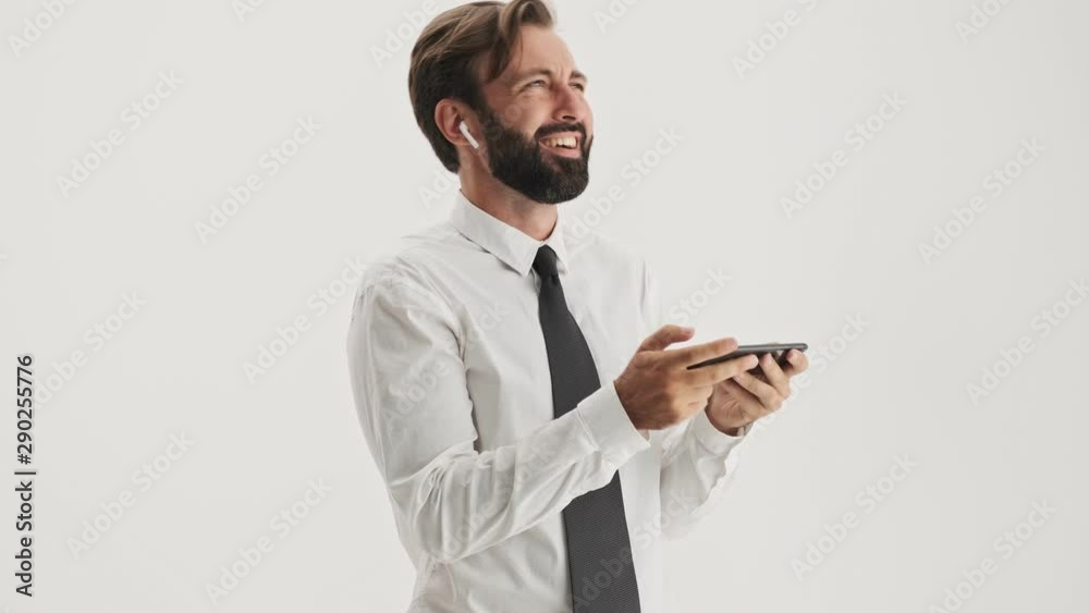 Cheerful young bearded business man with wireless earphones smiling and laughing while while watching video on smartphone over gray background isolated Cheerful young bearded business man with wireless earphones smiling and laughing while while watching video on smartphone over gray background isolated