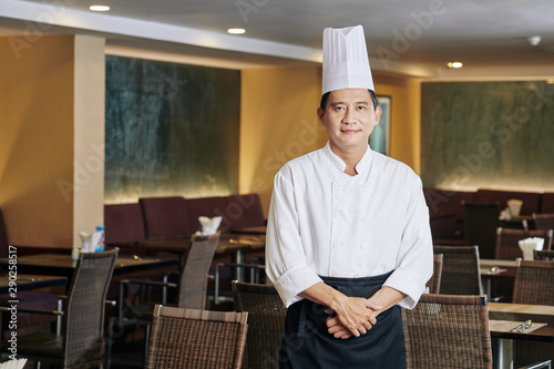 Portrait of Asian mature chef in uniform and in hat looking at camera while standing at empty hall of the restaurant