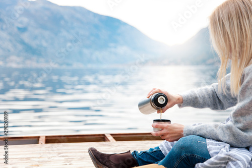 Cozy winter picnic on wooden pier by sea, blue mountains. Woman is pouring coffee from thermos into mug. Girl drinks hot steamy tea on beach. Girl is enjoying nature, relaxation, Christmas mood.