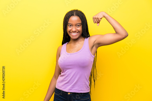 African American teenager girl with long braided hair over isolated yellow wall doing strong gesture