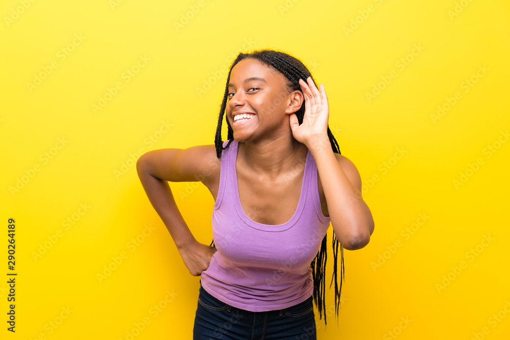 African American teenager girl with long braided hair over isolated yellow wall listening to something by putting hand on the ear