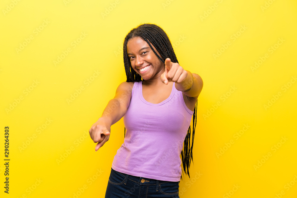 African American teenager girl with long braided hair over isolated yellow wall points finger at you while smiling African American teenager girl with long braided hair over isolated yellow wall points finger at you while smiling