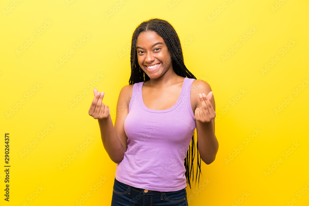 African American teenager girl with long braided hair over isolated yellow wall making money gesture African American teenager girl with long braided hair over isolated yellow wall making money gesture