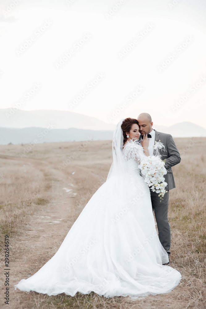 Just married loving couple in wedding dress and suit on field. Beautiful couple posing on the nature with bouquet of orchids.  Happy bride and groom walking and embracing in the summer meadow. 