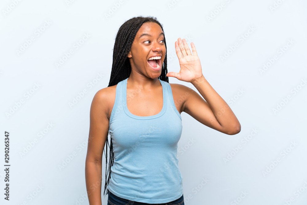 African American teenager girl with long braided hair over isolated blue background shouting with mouth wide open African American teenager girl with long braided hair over isolated blue background shouting with mouth wide open