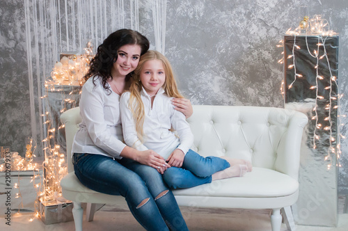 Two very pretty girls, sisters celebrates Christmas, new year. Girls in the New Year's interior of the room are sitting on a white sofa against the background of garlands