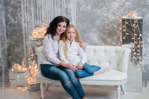 Two very pretty girls, sisters celebrates Christmas, new year. Girls in the New Year's interior of the room are sitting on a white sofa against the background of garlands