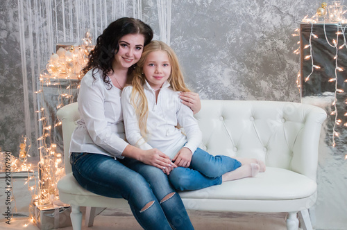 Two very pretty girls, sisters celebrates Christmas, new year. Girls in the New Year's interior of the room are sitting on a white sofa against the background of garlands