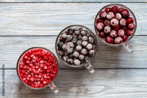 Ripe frozen sweet cherries, red currant and black currant with hoarfrost in the transparent glass cups on blue wooden background. Natural organic healthy food. Top view photo