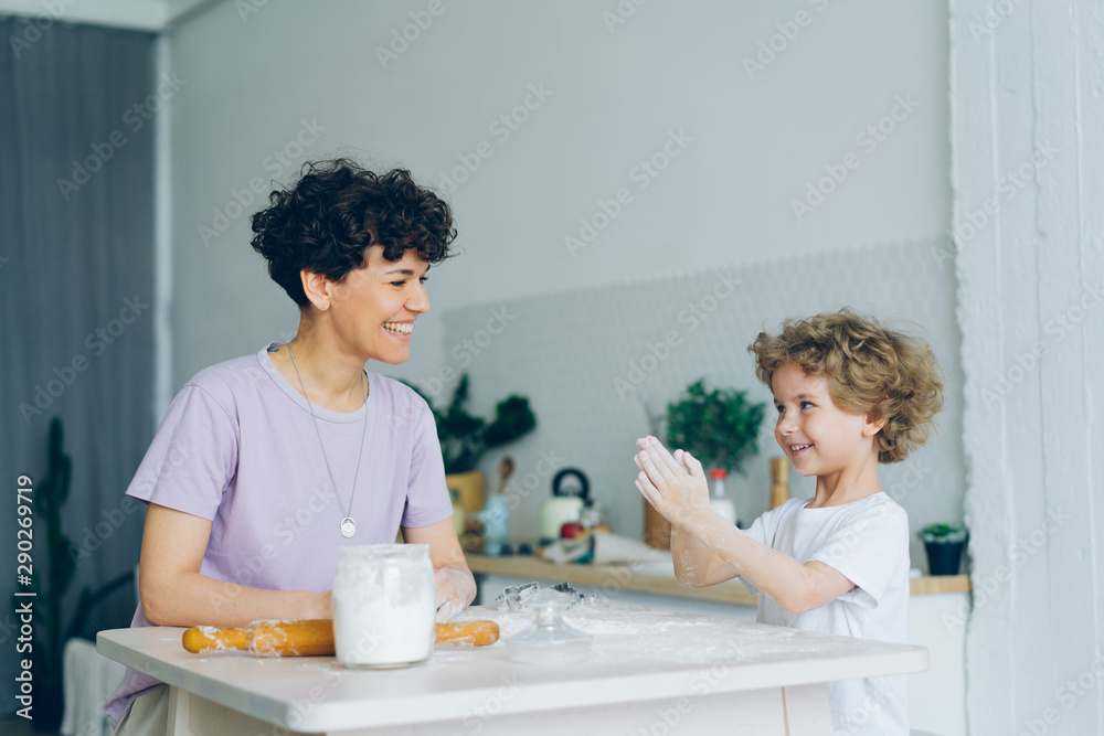 Happy child is having fun with flour clapping hands cooking pastry with mom sitting at table in kitchen at home. Joyful childhood, food and people concept. Happy child is having fun with flour clapping hands cooking pastry with mom sitting at table in kitchen at home. Joyful childhood, food and people concept.