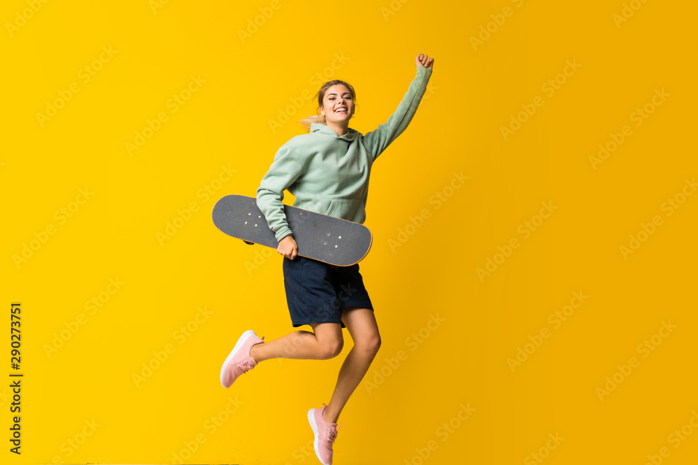 Blonde teenager skater girl jumping over isolated yellow background