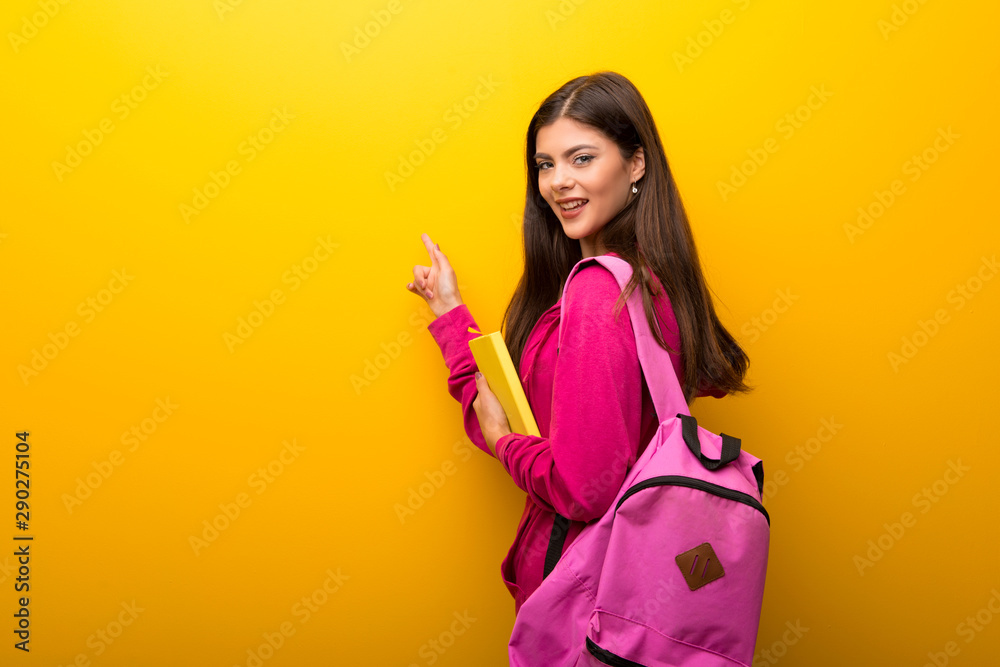 Teenager student girl on vibrant yellow background pointing back with the index finger Teenager student girl on vibrant yellow background pointing back with the index finger