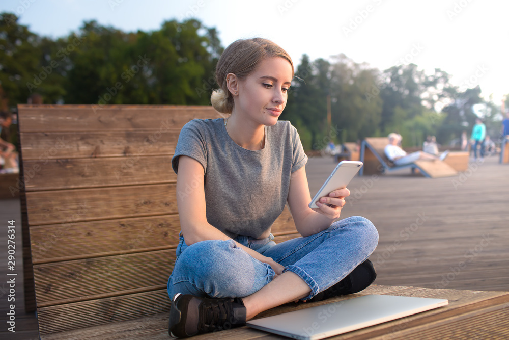 Stylish hipster girl using apps on cellphone while sitting with netbook on bench in park during summer vacation. Female blogger reading article on mobile phone