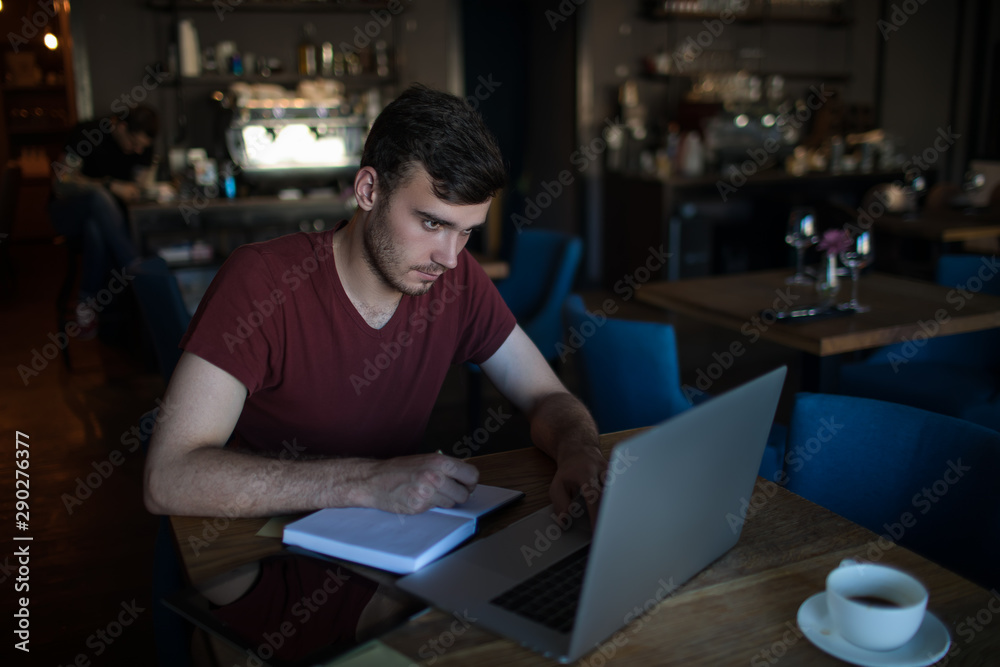 Man smart university student writing information in textbook from pc laptop computer, while sitting in coffee shop. Male content writer using netbook and diary for creating blog article