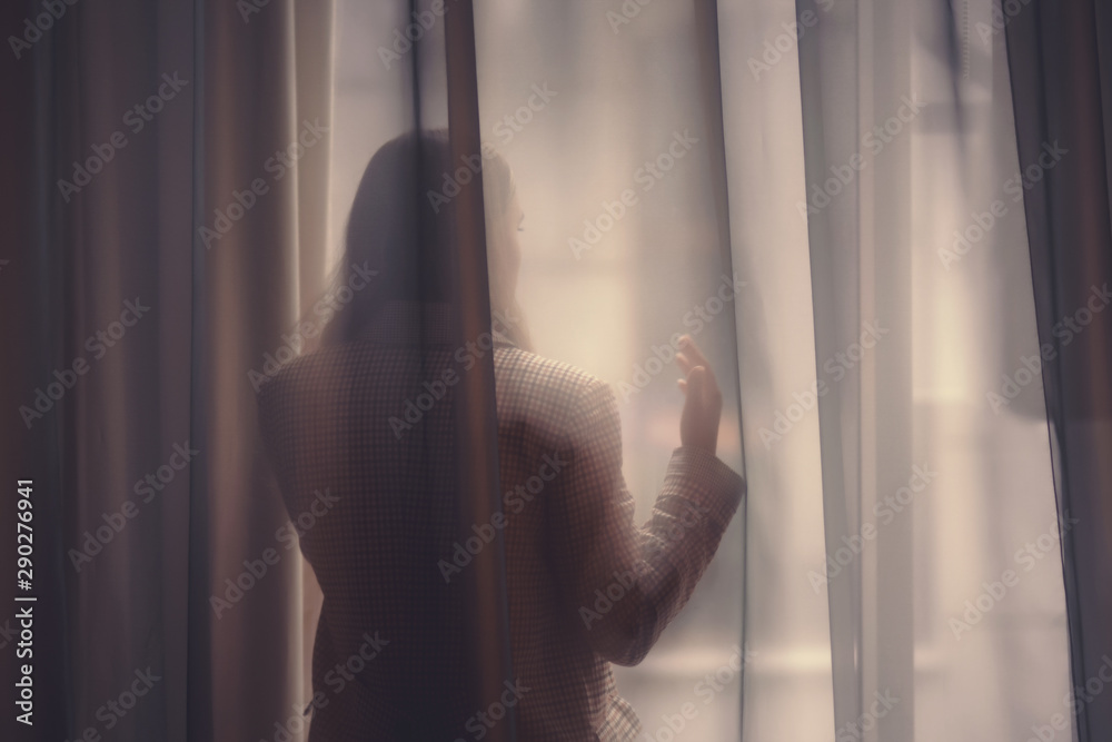 Moody shot of silhouette of a girl lean on the window. Beautiful girl stands behind the transparent curtain on the balcony early morning at sunrise.