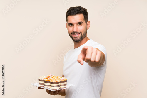 Handsome man holding muffin cake over isolated background points finger at you with a confident expression