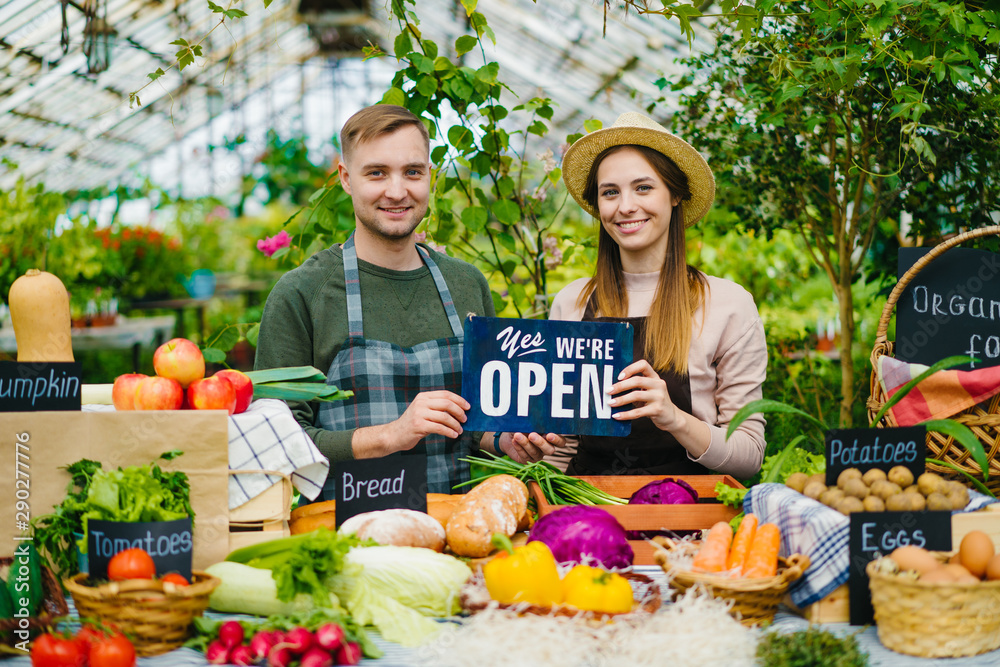 Guy and girl in aprons are holding open sign working at table with organic food in greenhouse market smiling looking at camera welcoming consumers.