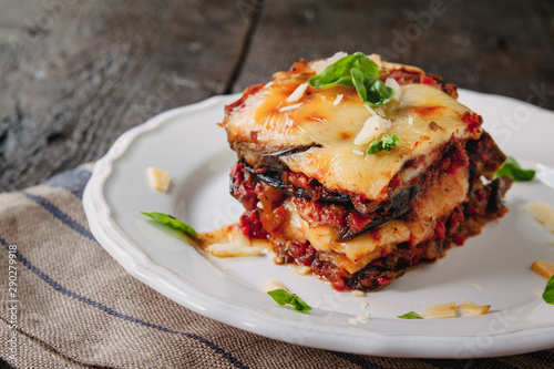 tradicional Parmigiana di melanzane: baked eggplant - italy, sicily cousine.Baked eggplant with cheese, tomatoes and spices on a white plate. A dish of eggplant is on a wooden table