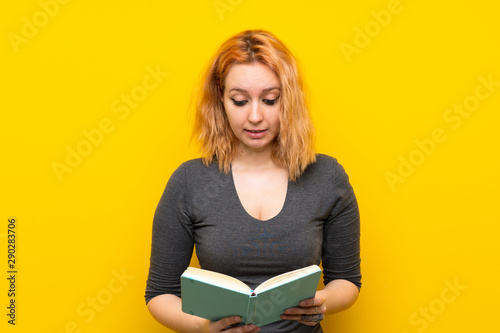 Young woman over isolated yellow background holding and reading a book