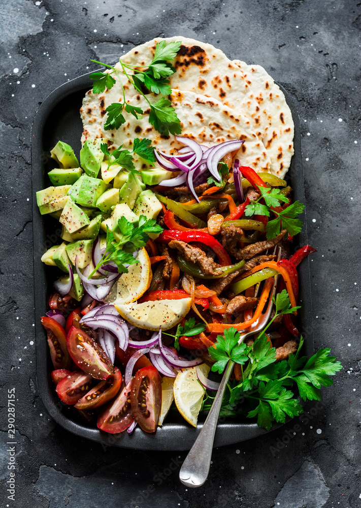 Spicy beef, vegetables, avocado, corn tortillas fajitas on a sheet pan on a dark background, top view. Delicious snack, tapas in mexican style