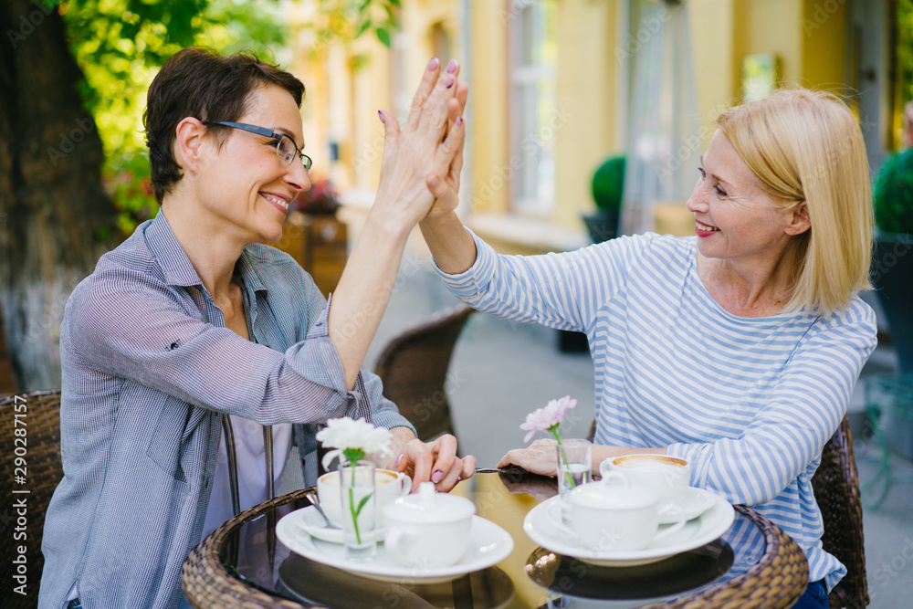 Joyful ladies friends are chatting doing high-five expressing unity and understanding sitting in outdoor cafe in city street. People, lifestyle and friendship concept. Joyful ladies friends are chatting doing high-five expressing unity and understanding sitting in outdoor cafe in city street. People, lifestyle and friendship concept.