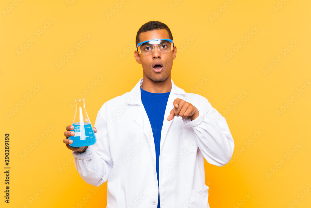 Scientific man holding a laboratory flask over isolated background surprised and pointing front Scientific man holding a laboratory flask over isolated background surprised and pointing front