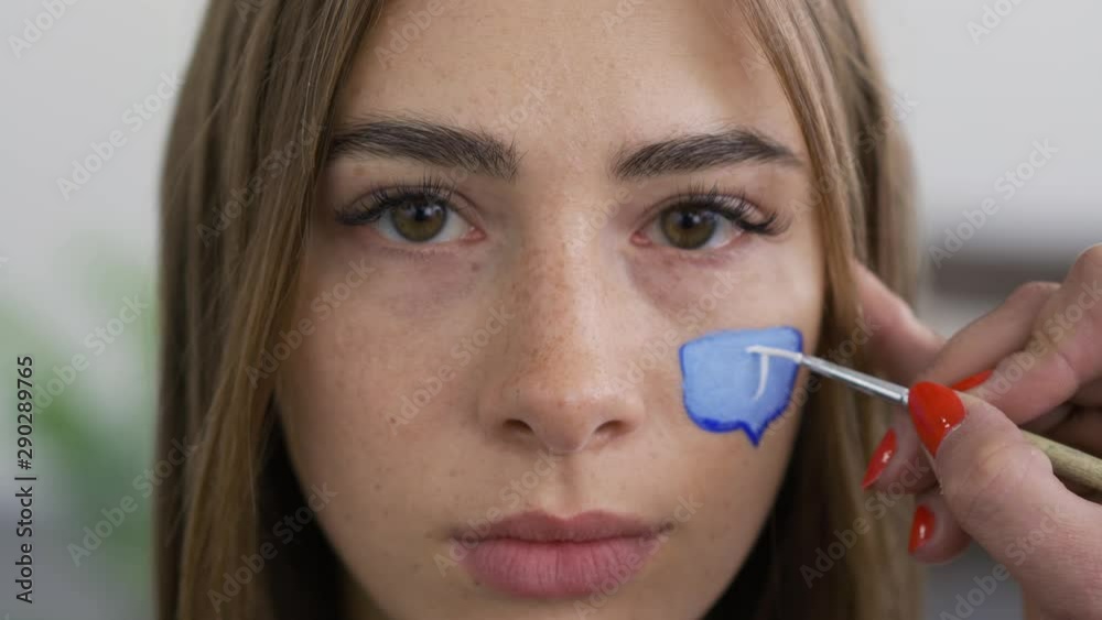 Close-up portrait of young pleasant woman looking at camera while artist painting zero on the message box on the girl's face using small brush. Social media concept.