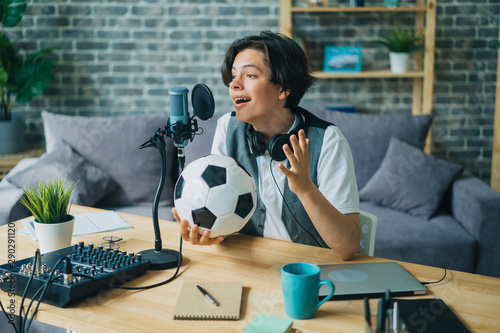 Joyful kid recording audio about football holding ball talking in microphone at home sitting at table alone. Youth lifestyle, sports fans and technology concept.