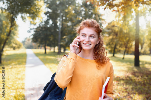 Smiling happy red hair student girl talking on the phone outside in autumn park