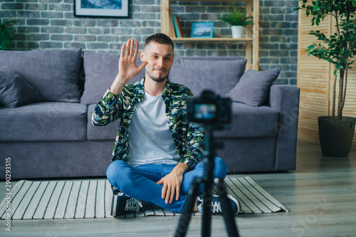 Handsome young man blogger is recording video for internet vlog talking smiling waving hand using camera on tripod sitting on floor in studio apartment. People and lifestyle concept. photo