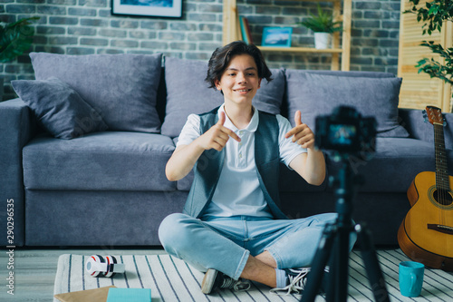Handsome teenager is showing thumbs-up recording video for internet vlog at home creating content for online blog. People, blogging and lifestyle concept. © silverkblack