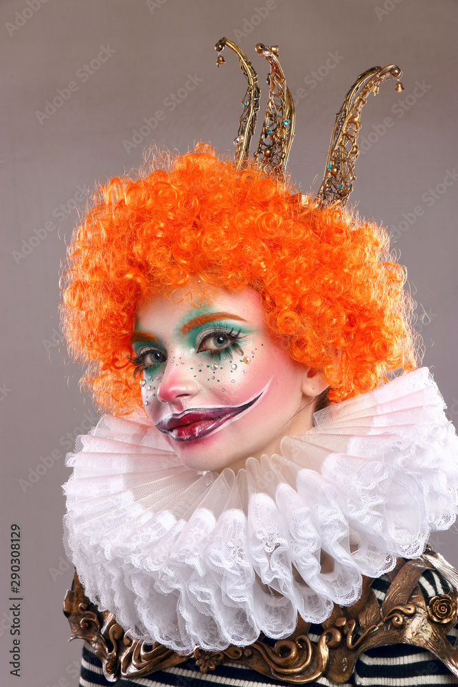 Red sad clown . Beautiful girl with bright makeup with wig and painted smile. vertical frame, gray background . Stock Photo Adobe Stock