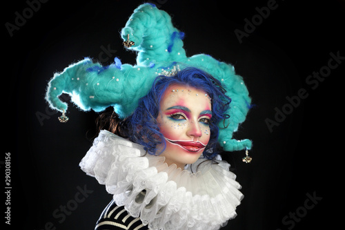 girl in makeup and costume jester . clown girl with bright makeup in blue wig On black background. he looks haughtily at the camera 
