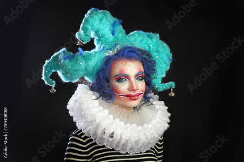 girl in makeup and costume jester . the clown girl is smiling, with bright makeup in a blue wig On a black background. he looks at the camera .