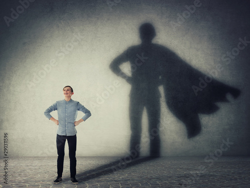 Fototapeta Casual teenage, keeps arms on hips smiling confident, casting a superhero with cape shadow on the wall
