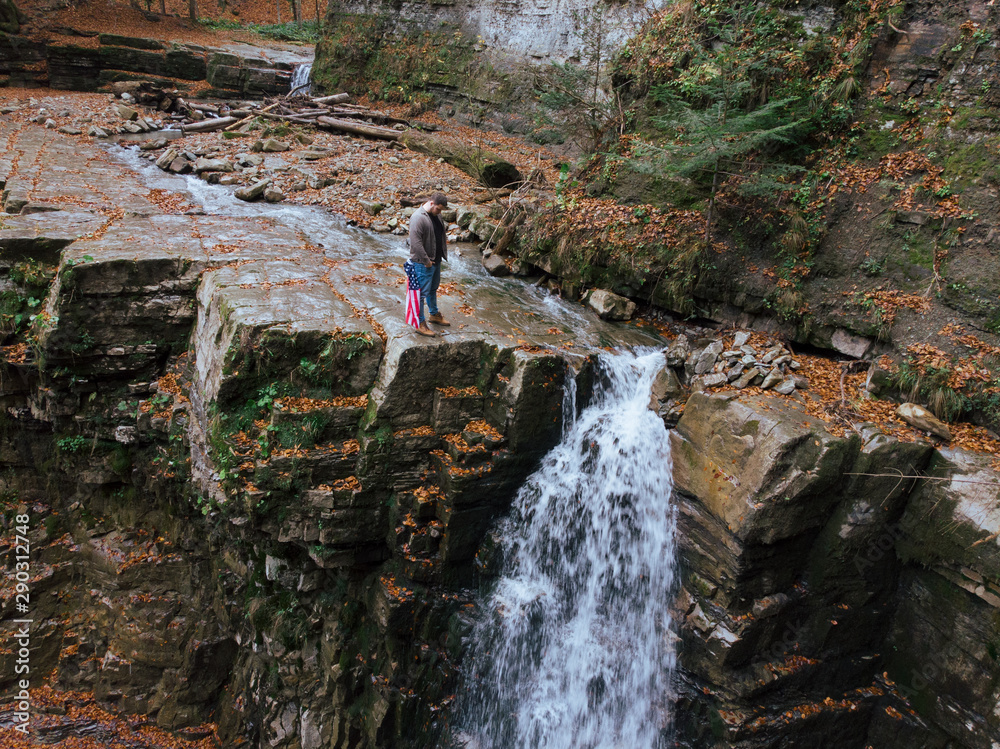 Fototapeta premium man holding usa flag on the top of the cliff autumn waterfall