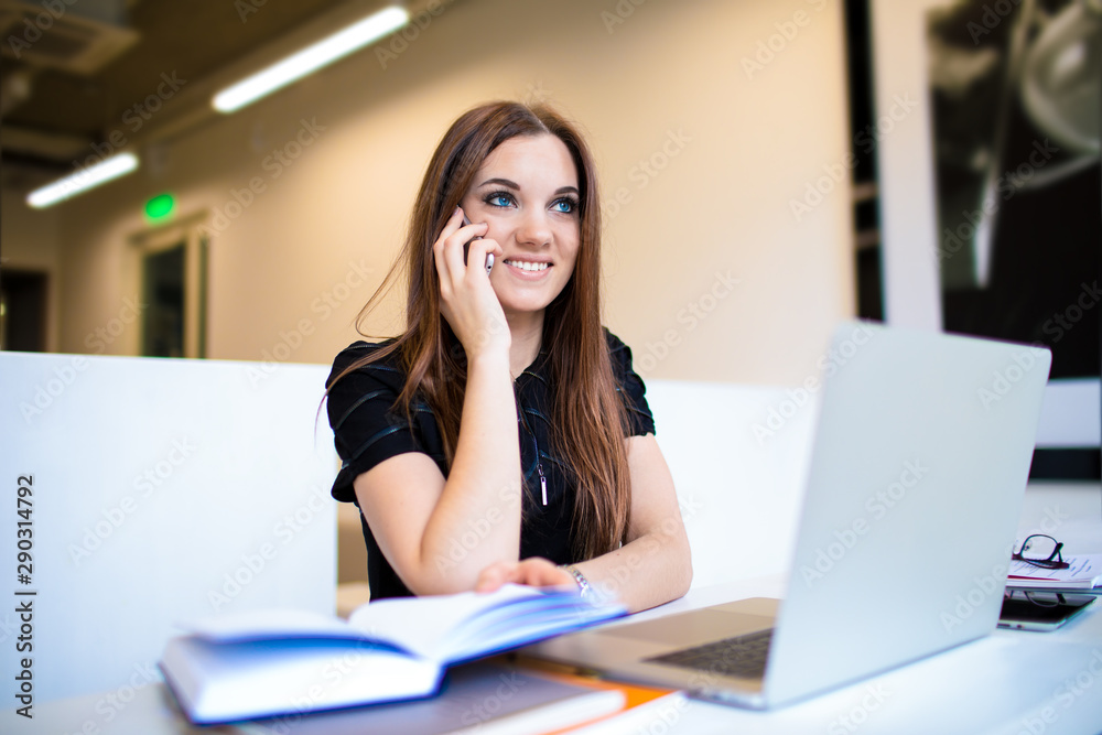 Happy female copywriter talking on mobile phone with friend during distance work on laptop computer, sitting in coworking space. Smiling female student having cellphone conversation during learning