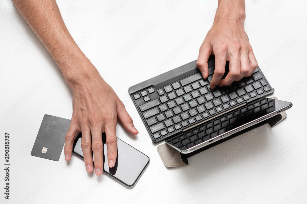 Close up top view of a man working on a project using digital tablet with keyboard seating at white table. Smartphone and bank card is near the tablet. Modern technologies at work. Close up top view of a man working on a project using digital tablet with keyboard seating at white table. Smartphone and bank card is near the tablet. Modern technologies at work.