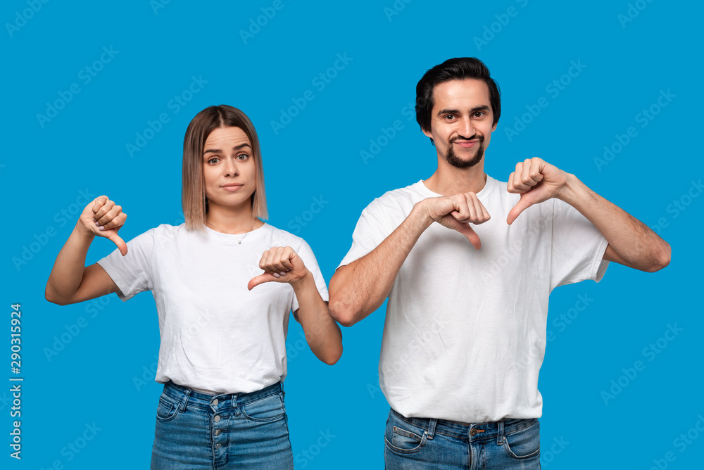 Upset young bearded man with mustaches and blond woman in white t-shirts and jeans holding thumbs down standing isolated over blue background. Concept of negative evaluation. Don't like it. Upset young bearded man with mustaches and blond woman in white t-shirts and jeans holding thumbs down standing isolated over blue background. Concept of negative evaluation. Don't like it.