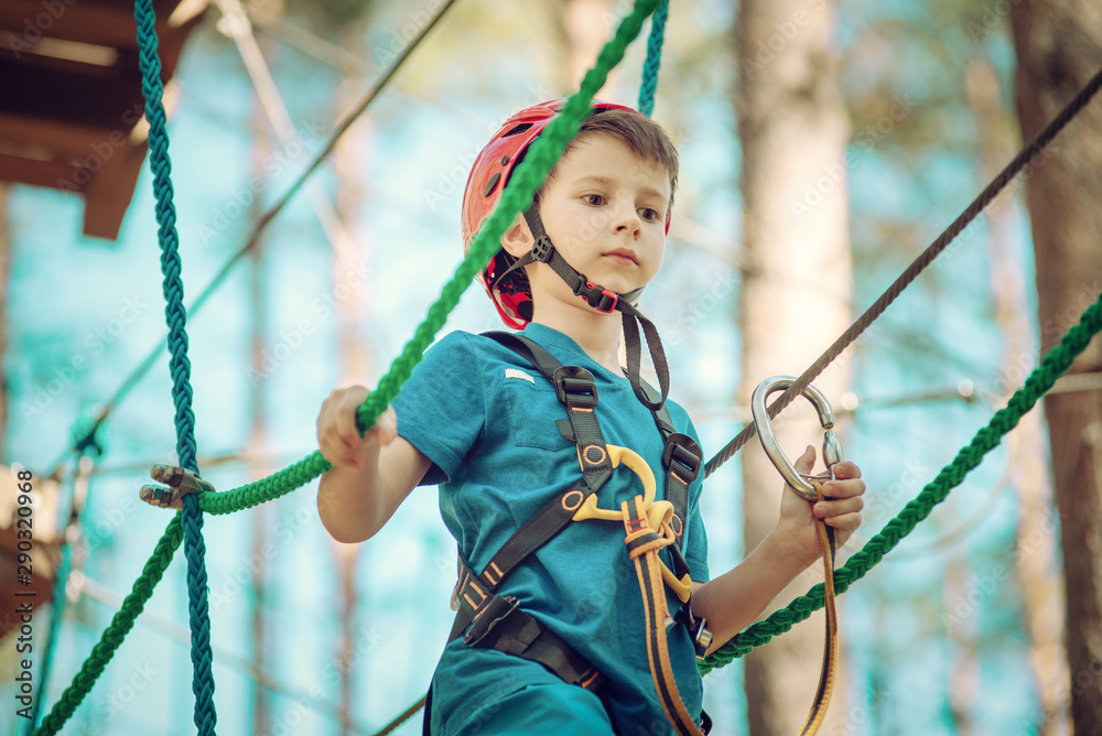 Boy enjoying activity in climbing adventure park at sunny summer day. Kid climbing in rope playground structure. Safe climbing with helmet insurance. Child in forest adventure park, extreme sport. Boy enjoying activity in climbing adventure park at sunny summer day. Kid climbing in rope playground structure. Safe climbing with helmet insurance. Child in forest adventure park, extreme sport.