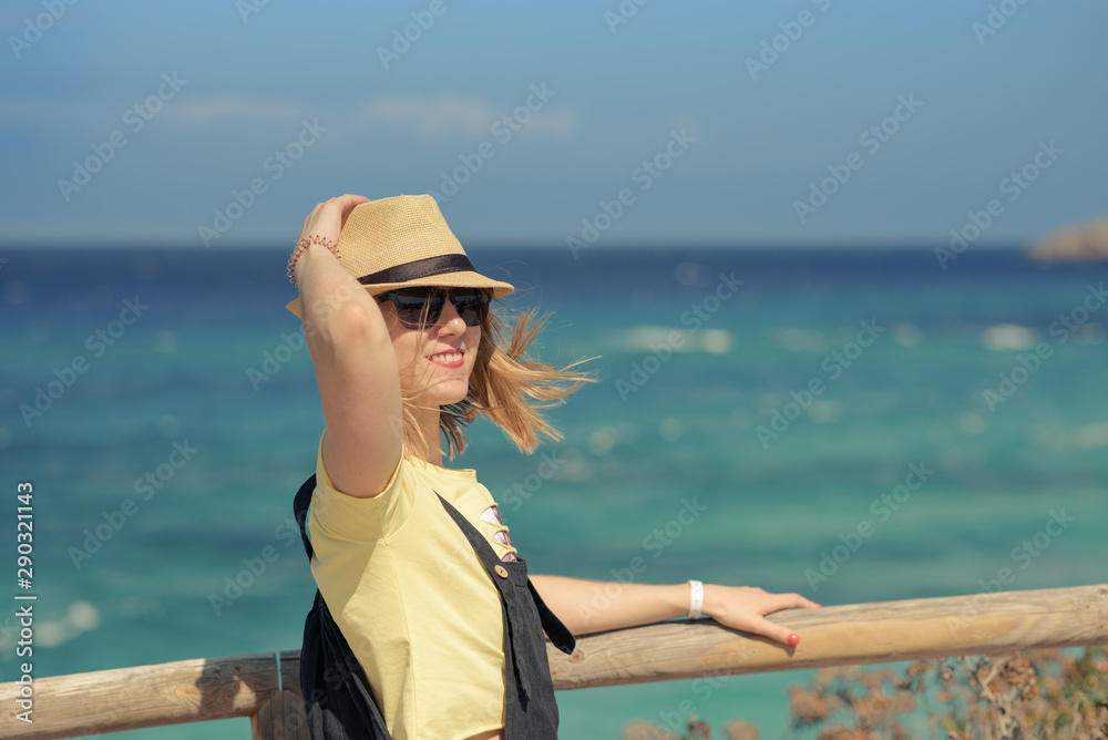 Smiling young European girl  in sun hat and sunglasses is standing on mountain beyond wonderful blue sea. She is having her best holidays ever on the amazing sea cost.