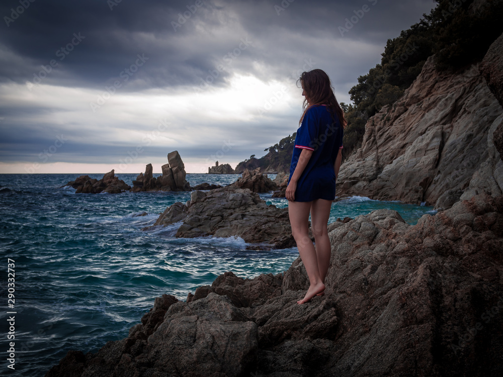 Caucasian brunette girl standing on the rocks near the rippling sea under dramatic skies in the twilight