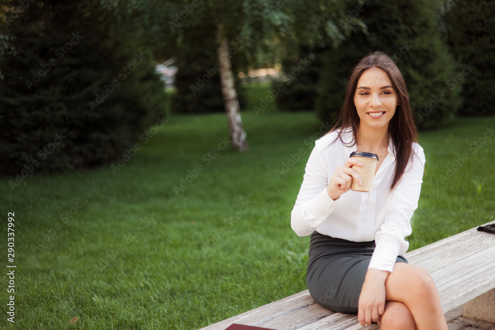 Young cheerful woman manager drink coffee while sitting on bench in park. Business concept Young cheerful woman manager drink coffee while sitting on bench in park. Business concept